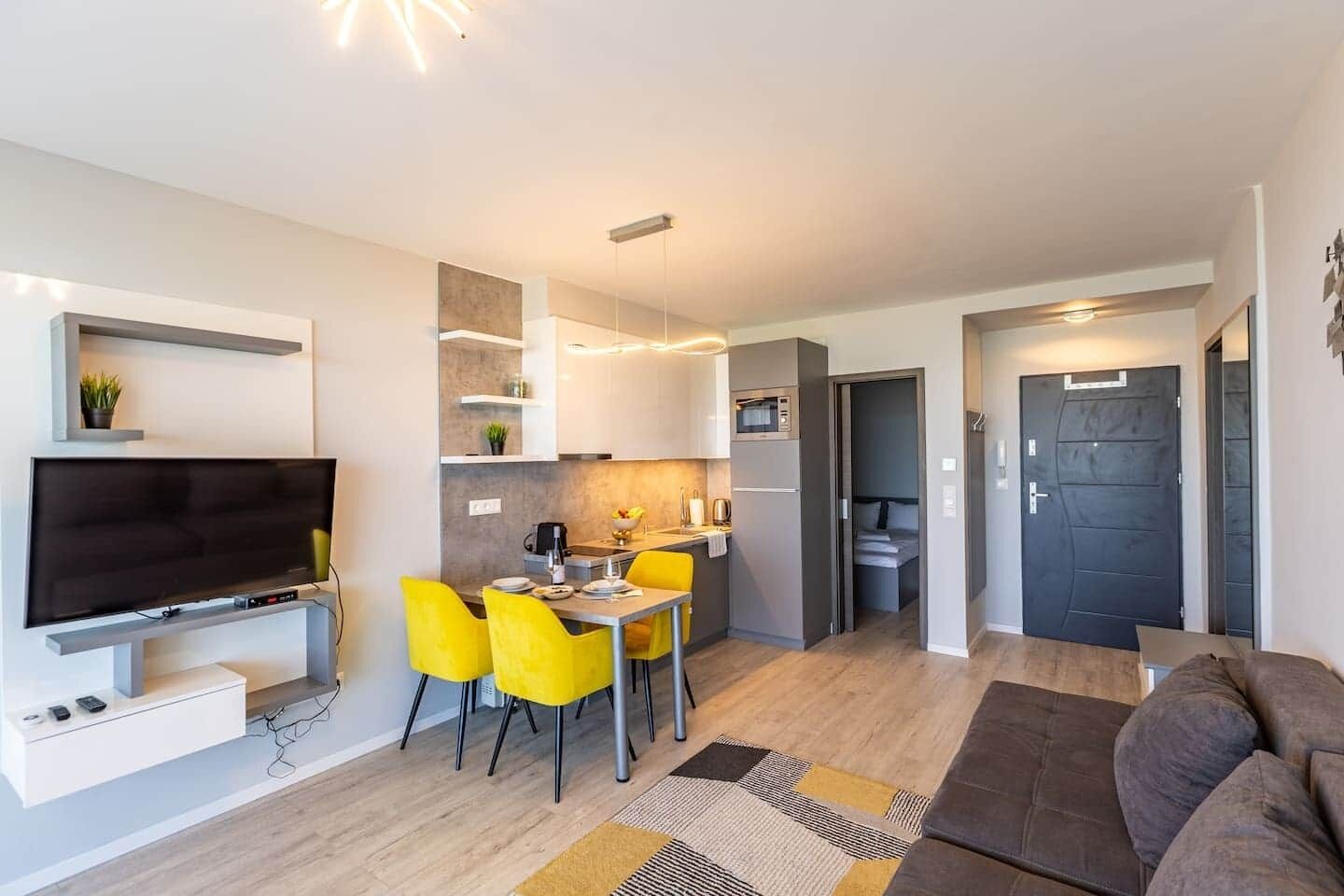 A compact living space with a flat-screen TV, a dining area with yellow chairs, a kitchenette, and a dark door.