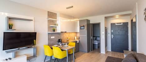 A compact living space with a flat-screen TV, a dining area with yellow chairs, a kitchenette, and a dark door.