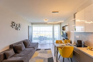 A modern living room with a sofa, dining table, and chairs, a kitchen with cabinets and appliances, and a large window with a view. - LB - LelleMarine Apartments A002 (Balatonlelle)