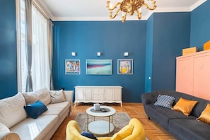 A living room with a blue wall, a white sofa, a black sofa, a yellow armchair, a white cabinet, and a chandelier. - MidTerm - Chic Soho Apartment near Opera (Budapest)