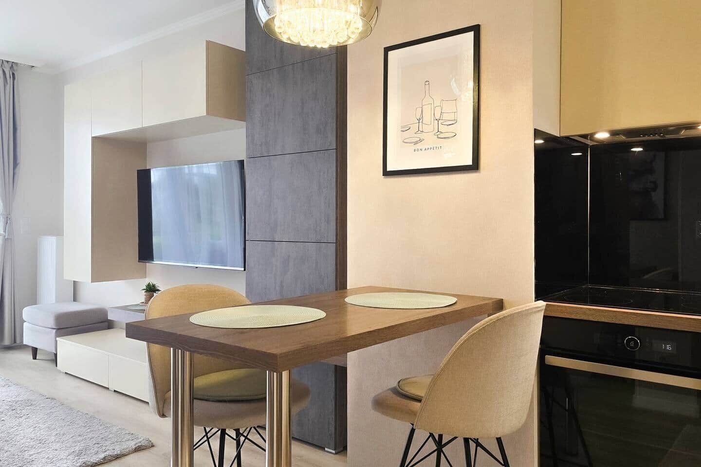 A modern living room with a dining area, a flat-screen TV mounted on the wall, a chandelier, and a framed artwork.