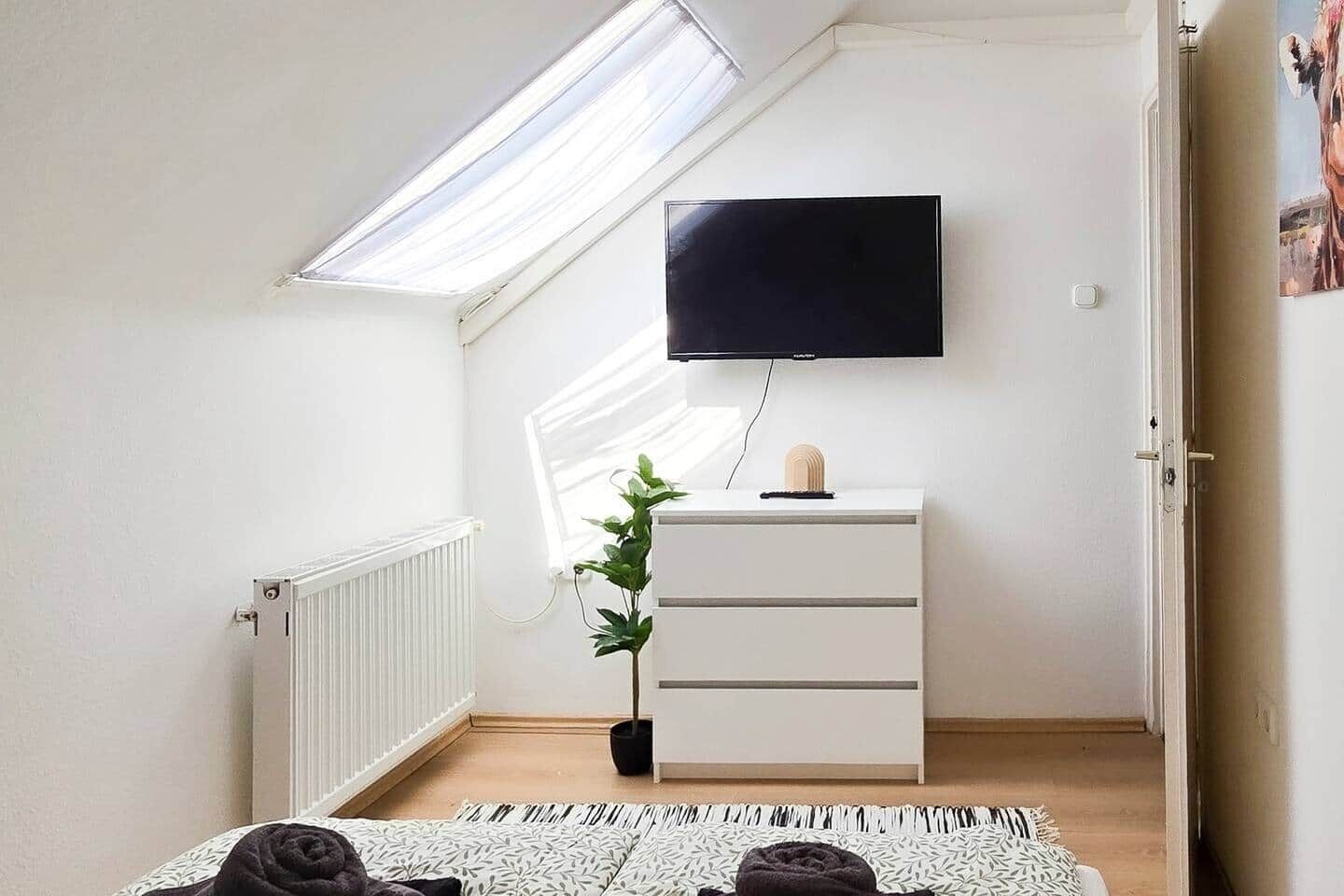 Una habitación con techo inclinado, un televisor de pantalla plana en la pared, un aparador blanco con cajones, una planta en maceta y una cama con ropa de cama estampada.