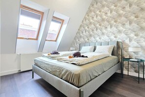 A modern bedroom with a sloped ceiling, a bed with a grey headboard, two windows with wooden frames, a radiator, and a geometric patterned wall. - MidTerm - Rooftop Serenity Apartment 3 (Budapest)