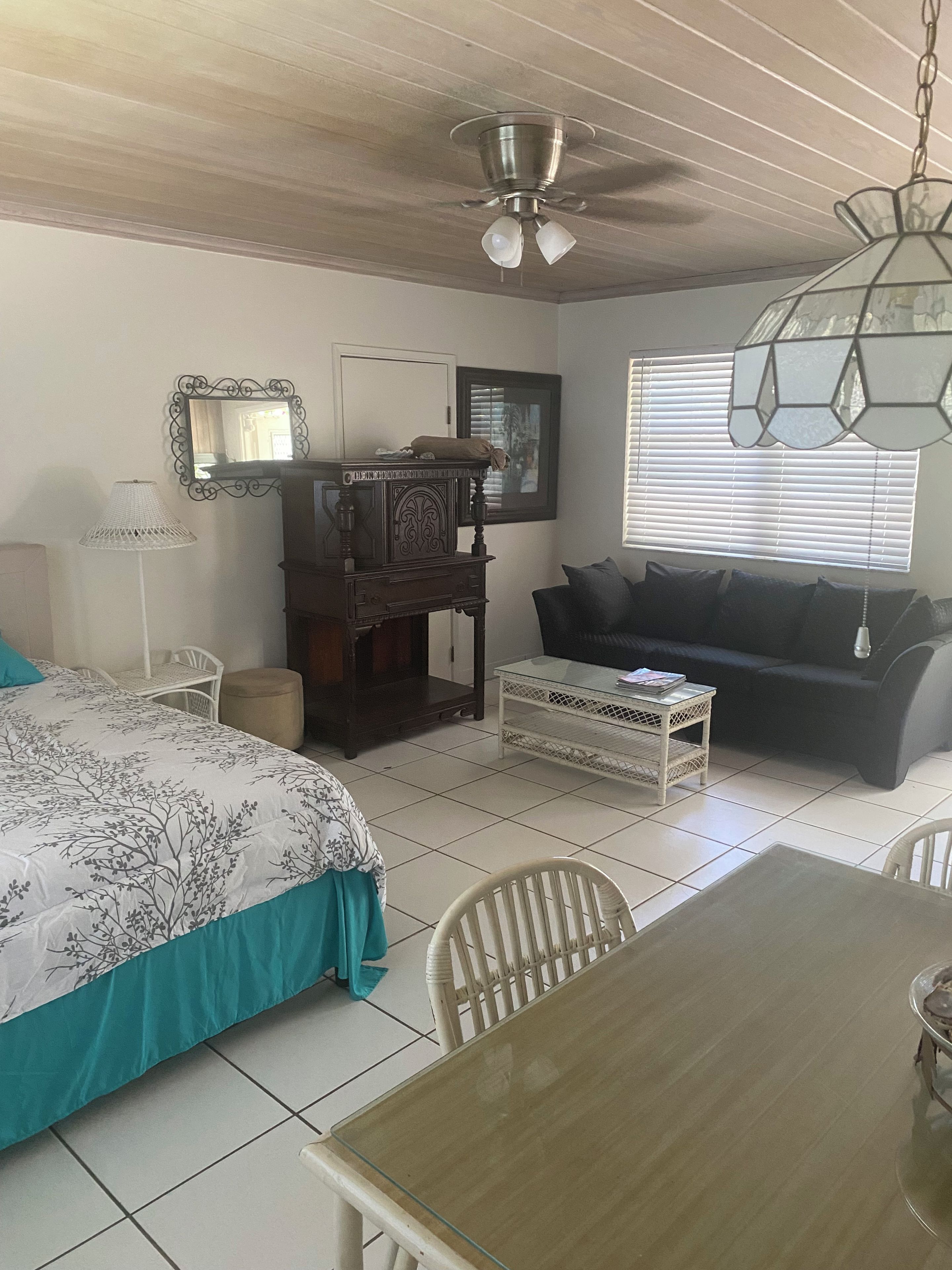 A room with a bed, a wooden table, a sofa, a chair, a lamp, a ceiling fan, and a window with blinds.