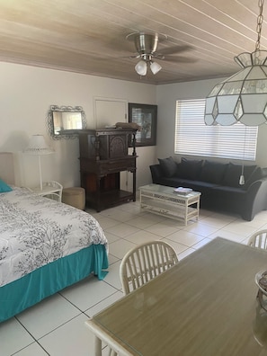 A room with a bed, a wooden table, a sofa, a chair, a lamp, a ceiling fan, and a window with blinds.