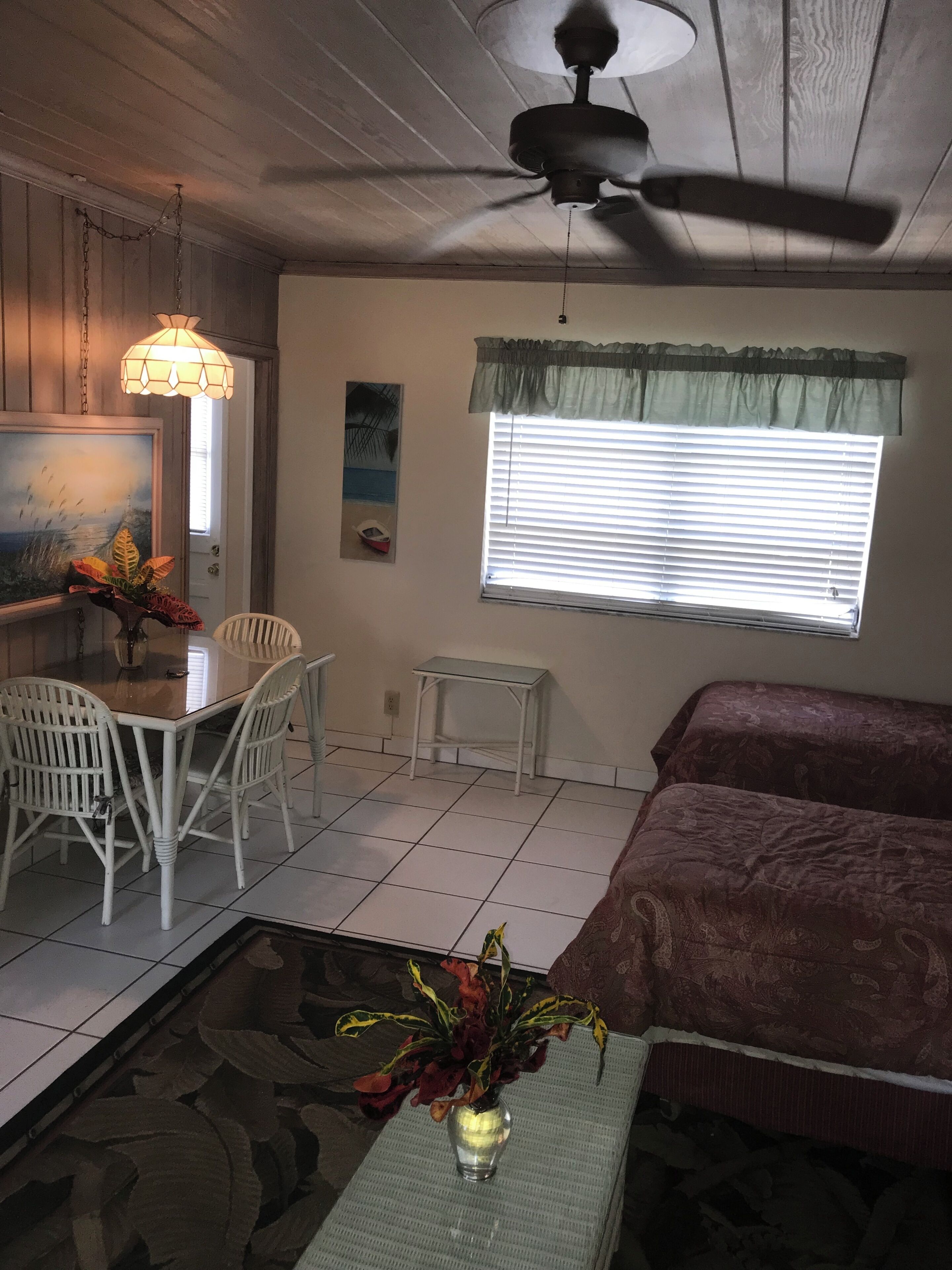 Ein Zimmer mit Esstisch, Stühlen, einem Bett, einem Fenster mit Jalousien, einem Deckenventilator und einer Vase mit Blumen.