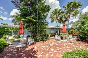A patio with outdoor furniture and umbrellas, surrounded by lush greenery and a building with a tiled roof.