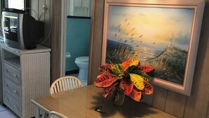 Et spiseområde med et træbord, hvide stole, en vase med blomster, et maleri og et fjernsyn på et stativ.