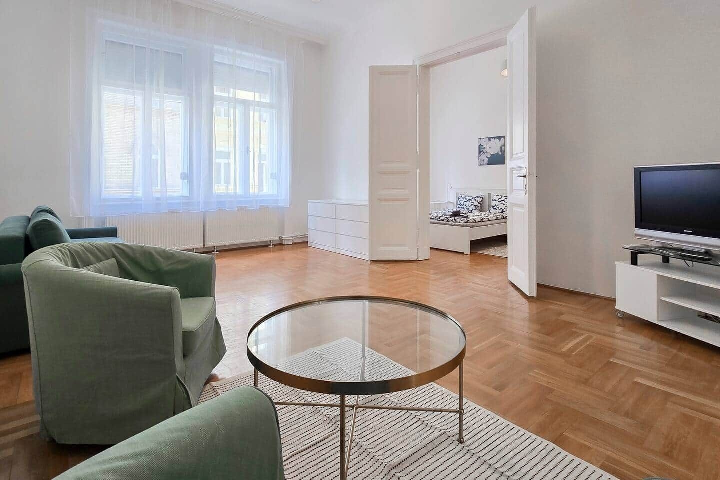 A modern living room with a glass coffee table, a sofa, a television on a stand, and a view through an open door.