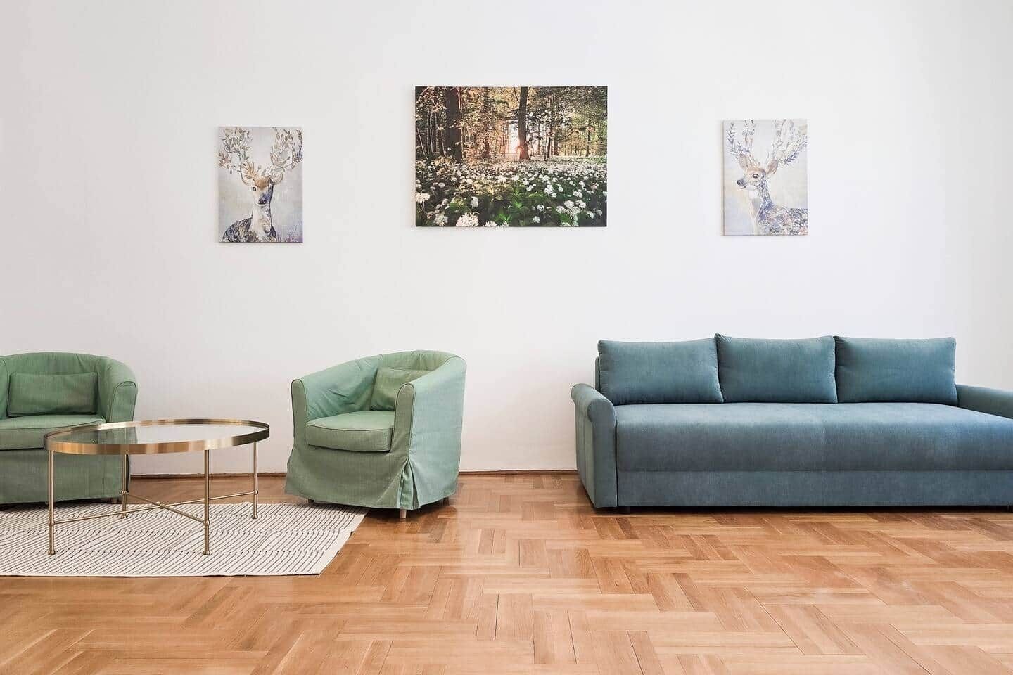 A living room with a green sofa, a blue sofa, two green armchairs, and a small round table. There are three framed pictures on the wall.