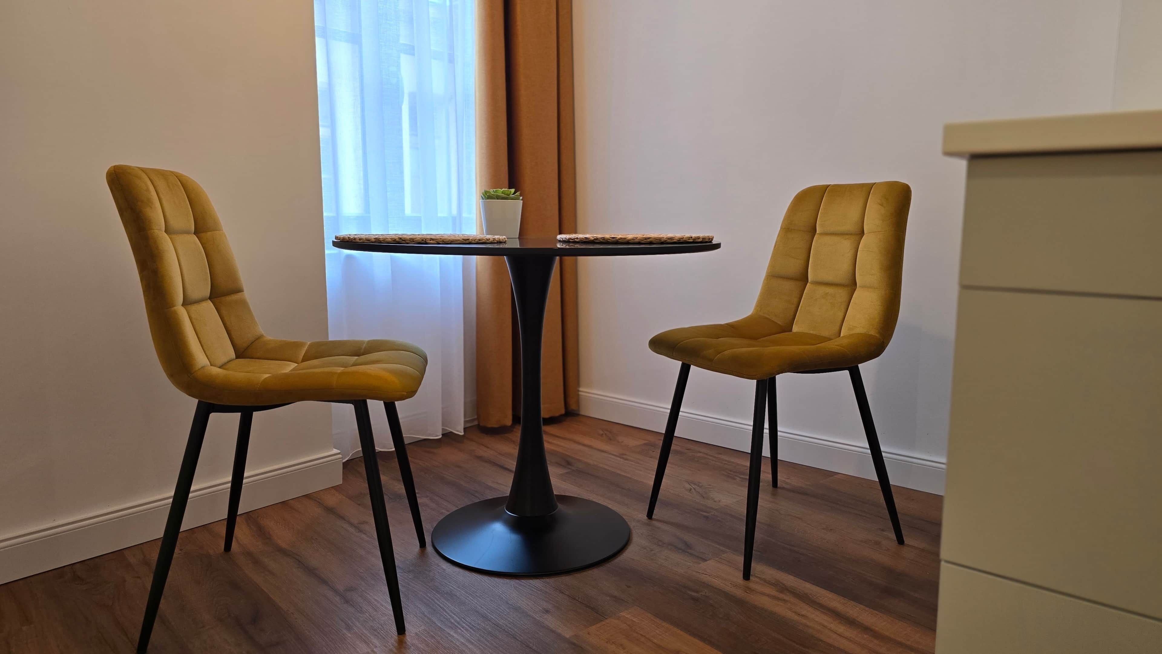 Two chairs with a round table in a room with wooden flooring and a window with curtains.
