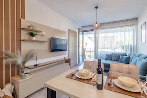 A modern living room with a flat-screen TV mounted on the wall, a dining table set with plates and wine glasses, and a sofa with cushions. - LB - LelleMarine Apartments B103 (Balatonlelle)
