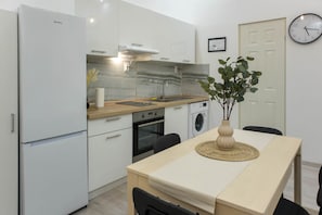 A compact kitchen with a refrigerator, oven, sink, and washing machine. There's a dining table with chairs and a vase with greenery. - MidTerm - Magpie Apartment (Budapest)