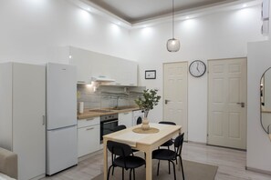 A modern kitchen with white cabinetry, a wooden countertop, and a built-in oven. There is a dining table with chairs and a vase with a plant. A clock is on the wall, and a door is visible in the background. - MidTerm - Magpie Apartment (Budapest)