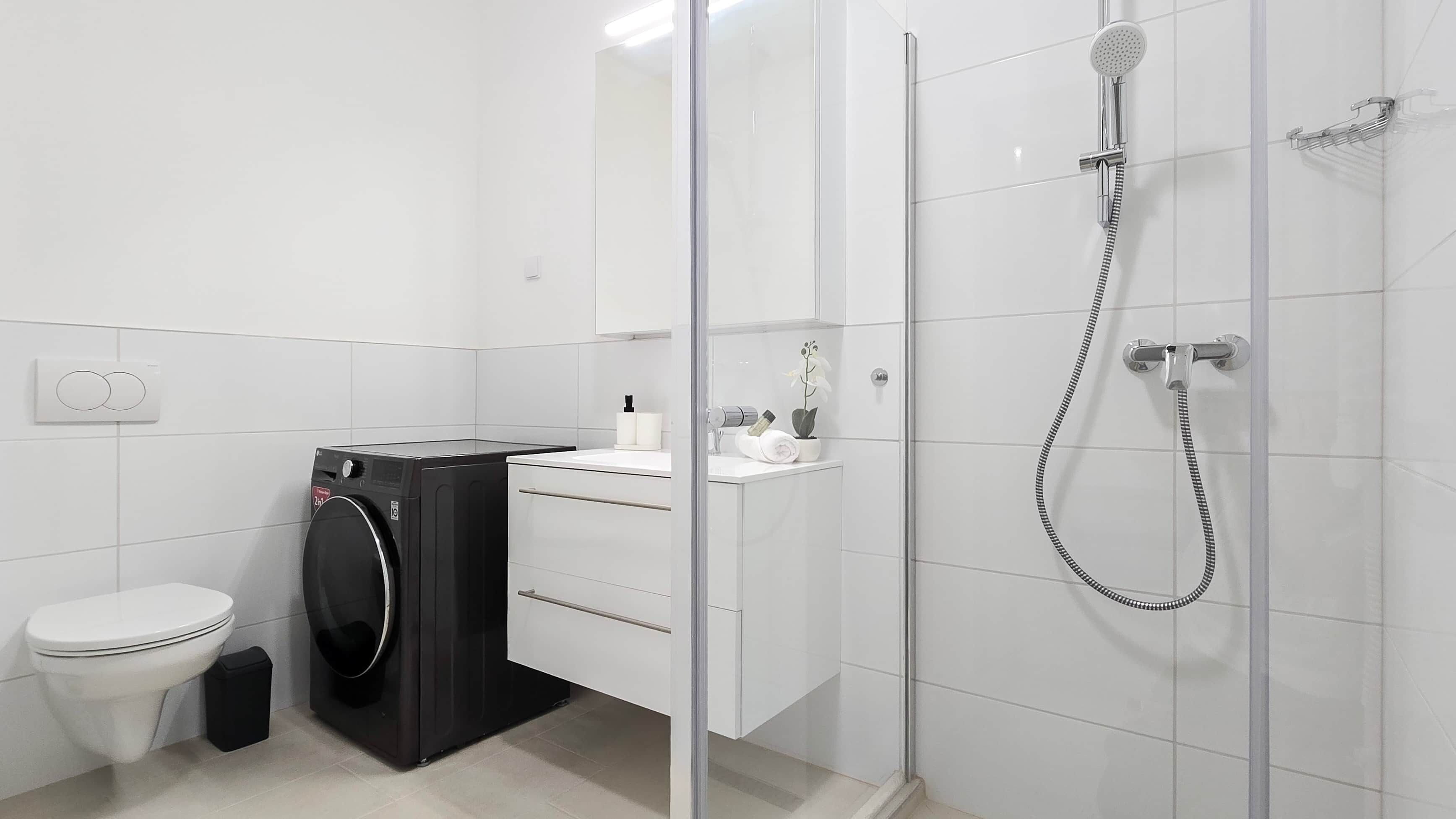 A modern bathroom with a glass-enclosed shower, a toilet, a washing machine, and a sink with toiletries.