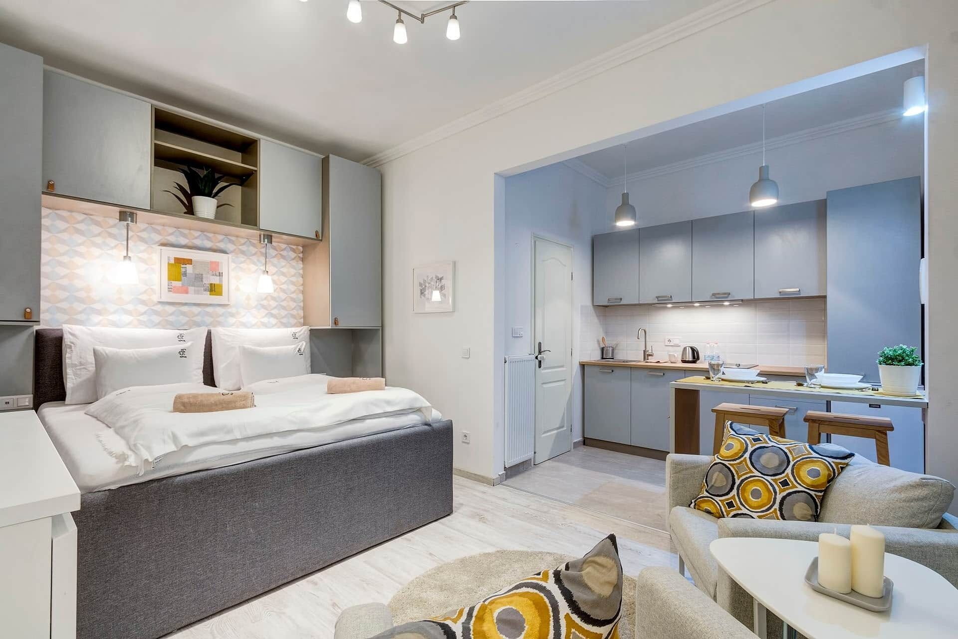 A modern hotel room with a bed, a sofa, a small table, and a kitchen area.