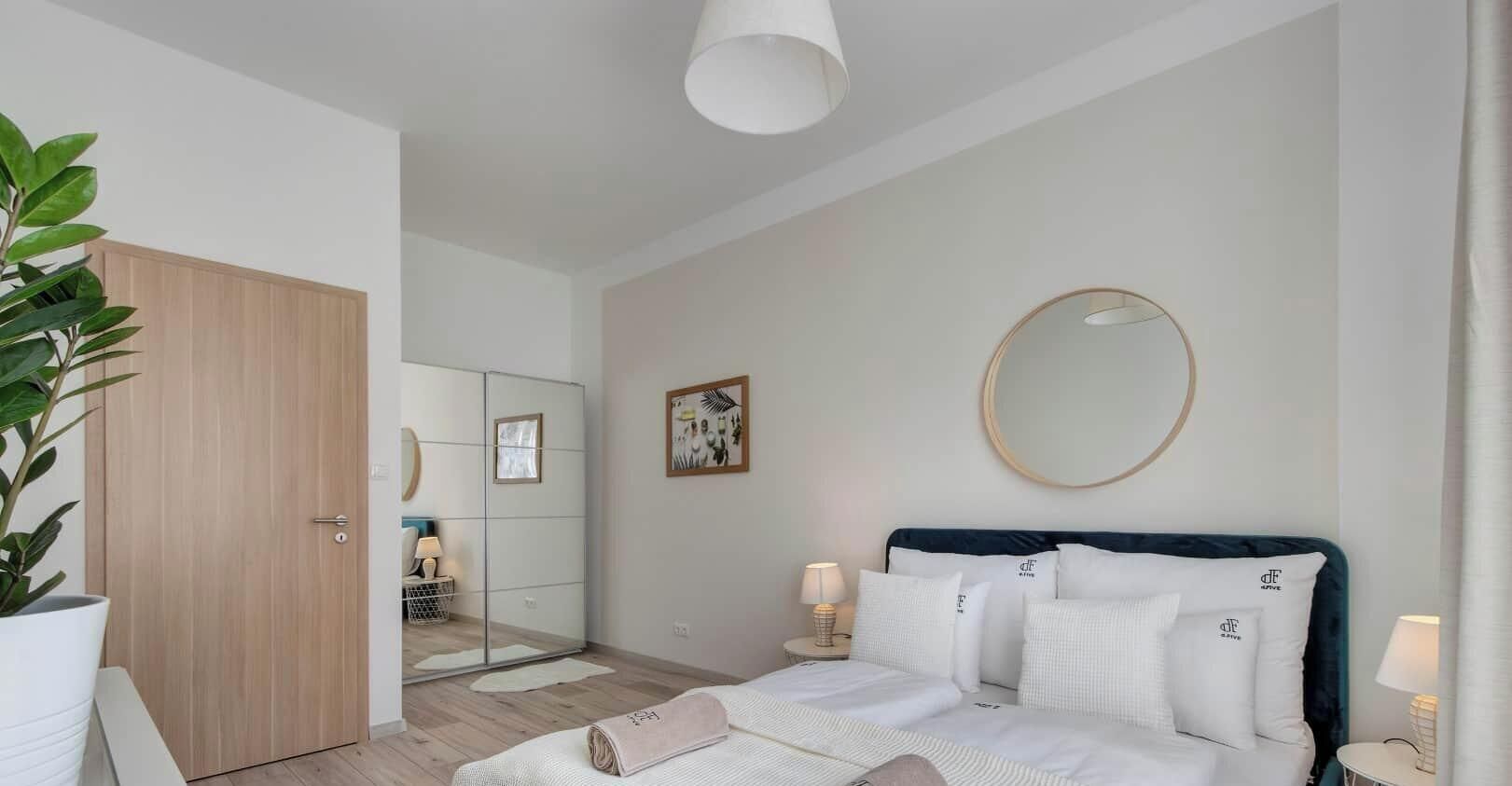 A hotel room with a bed, bedside lamps, a mirror, a potted plant, and a wooden wardrobe.