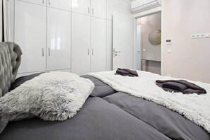 A bedroom with a bed, grey bedding, white fluffy pillows, a grey upholstered headboard, and white wardrobes. - LB - SunSet Panorama Apartment (Siófok)