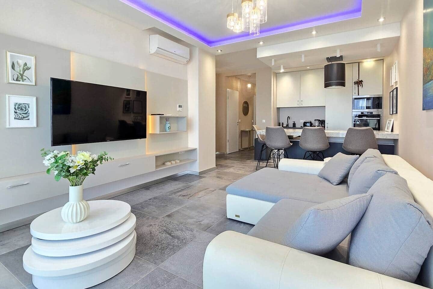 A modern living room with a flat-screen TV, a white console table, a grey sofa, and a dining area with chairs.