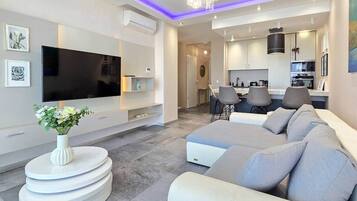 A modern living room with a flat-screen TV, a white console table, a grey sofa, and a dining area with chairs.