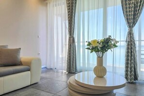 A living room with a sofa, a round table with a vase of flowers, and sheer curtains. - LB - SunSet Panorama Apartment (Siófok)