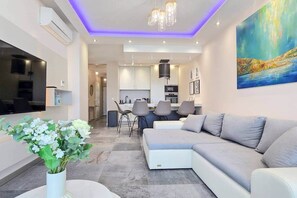 A modern living room with a sectional sofa, a dining area with chairs, a kitchen in the background, and a large painting on the wall. - LB - SunSet Panorama Apartment (Siófok)