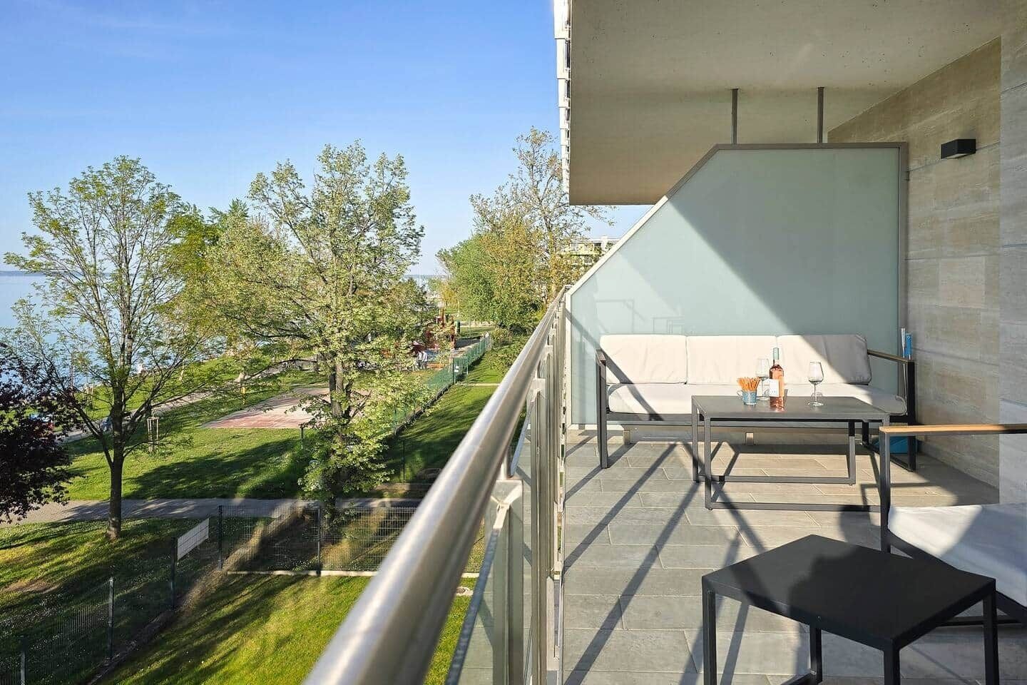 Un balcon avec un canapé et une table, donnant sur un parc arboré et sur un bâtiment situé au loin.