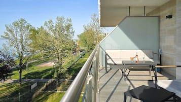 A balcony with a sofa and a table, overlooking a park with trees and a distant building.