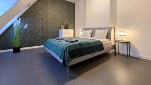 A modern bedroom with a geometric-patterned wall, a bed with a green blanket, a bedside table with a lamp, and a potted plant. - IMCT - Rooftop Serenity Apartment (Budapest)