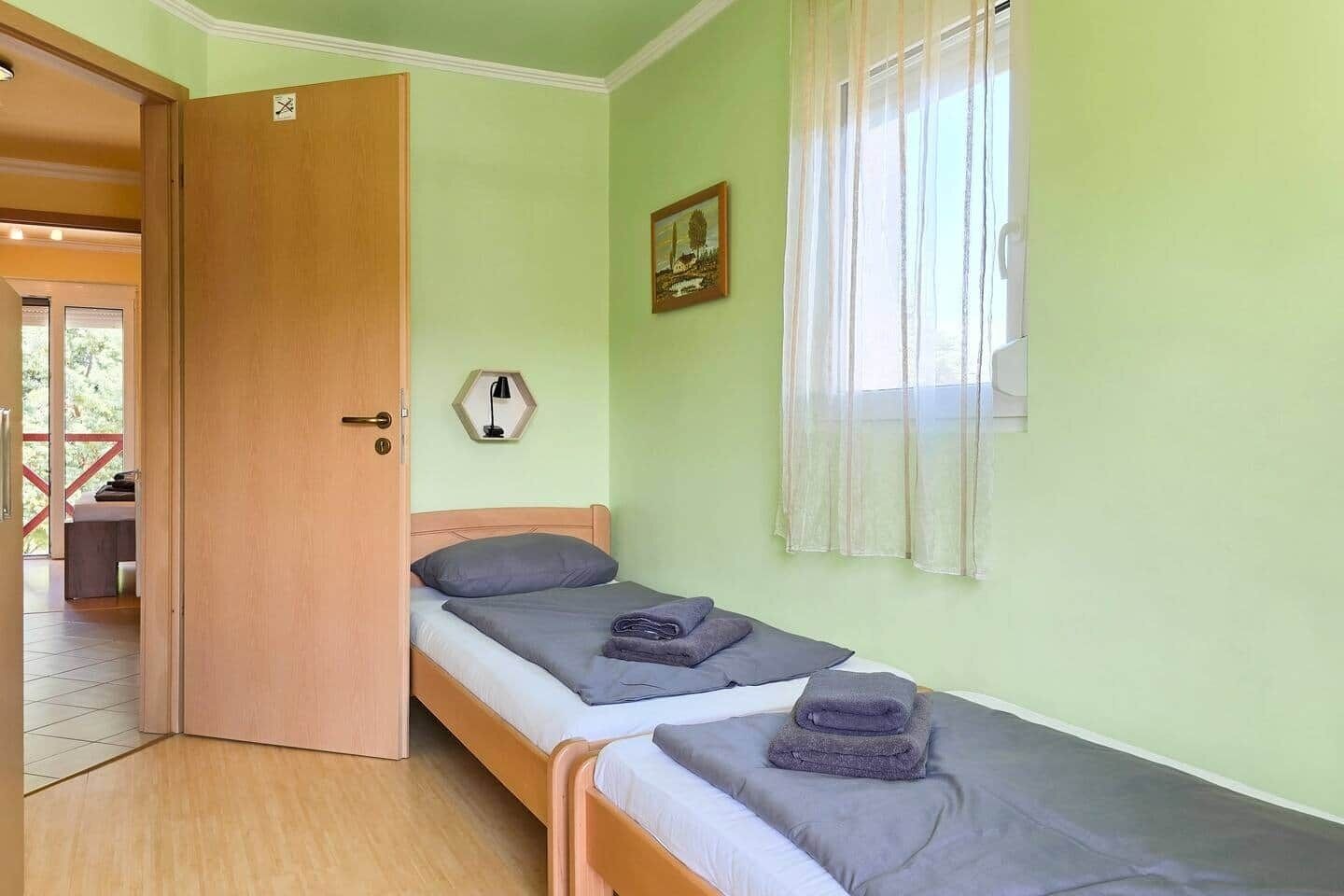 Um quarto com uma cama, uma porta, uma janela com cortinas e uma luminária fixada na parede.