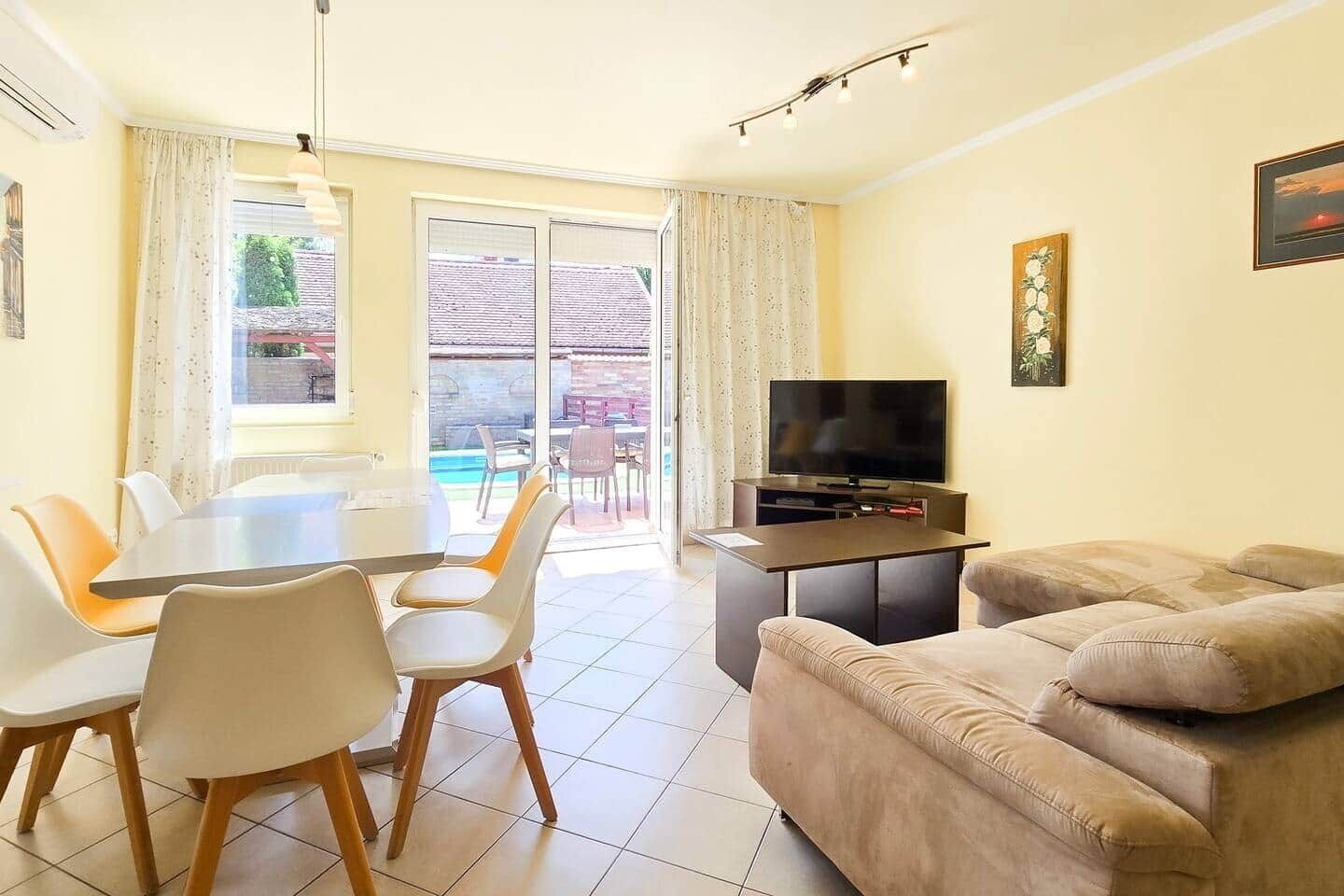 Un salon comprenant un canapé, un téléviseur, une table à manger avec des chaises et un balcon avec une piscine.