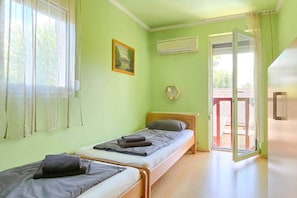 Ein Zimmer mit zwei Einzelbetten, einem Fenster mit durchsichtigen Vorhängen, einer Klimaanlage und einer zur Außenseite führenden Tür.