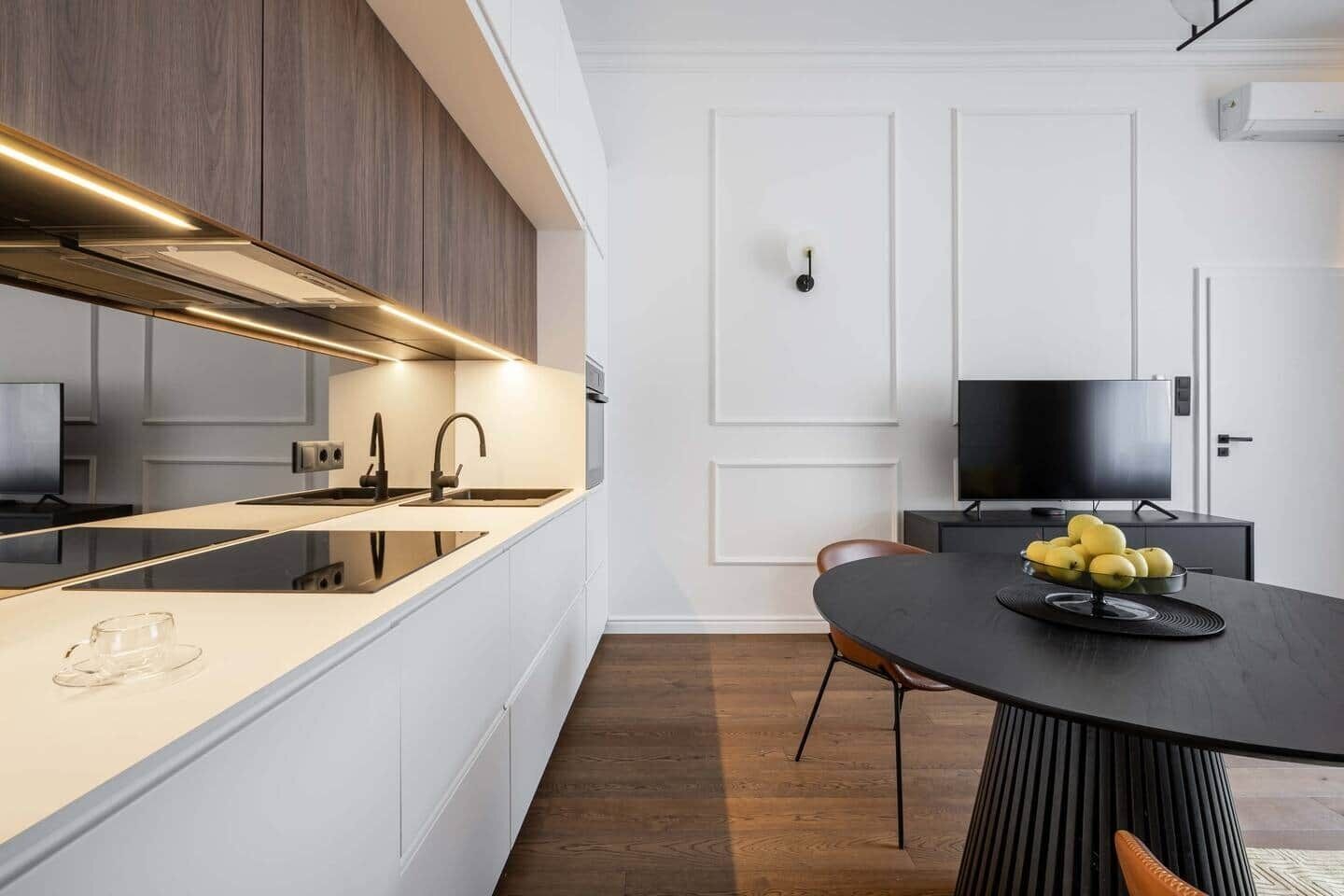 Una cocina moderna con horno integrado, una mesa de comedor con un bol de fruta y un televisor al fondo.