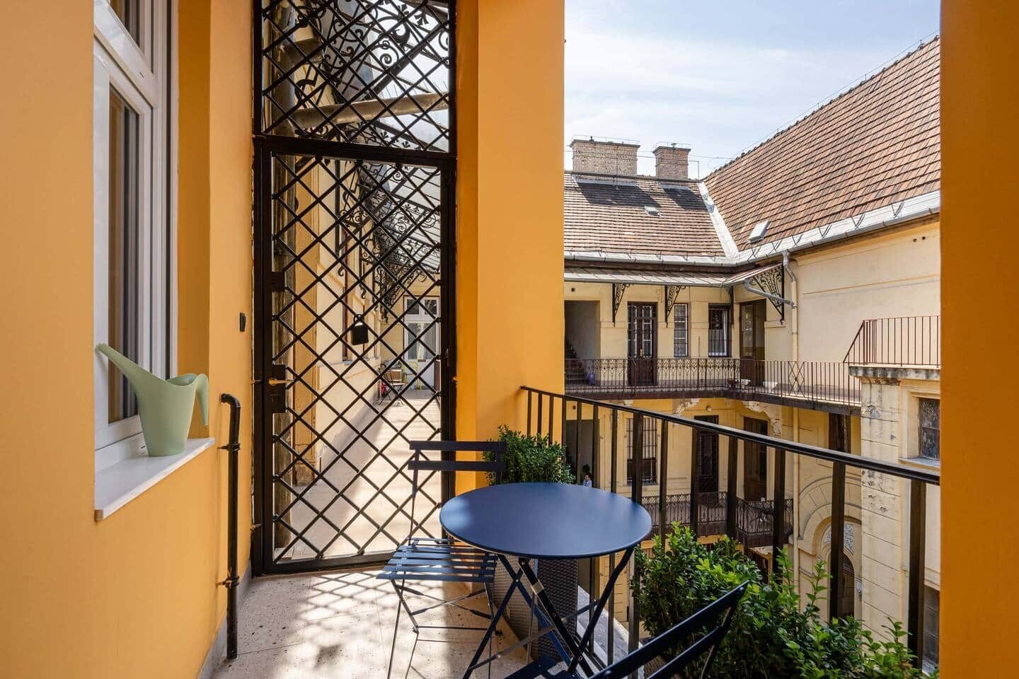 A balcony with a table and chairs, a gate, and a view of adjacent buildings.