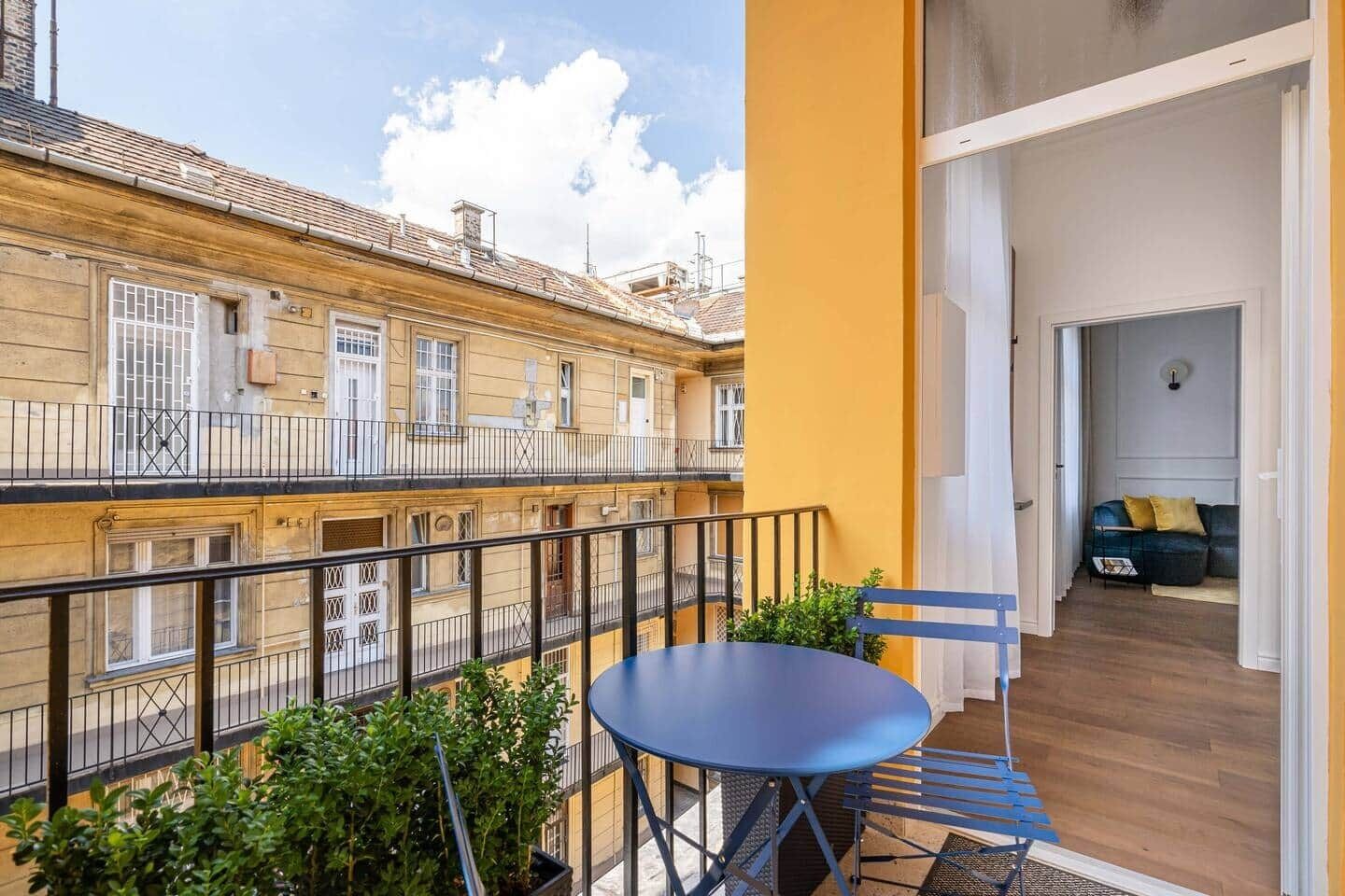 Un balcón con una mesa y sillas, vista de edificios vecinos y una puerta que da a un espacio interior con un sofá.