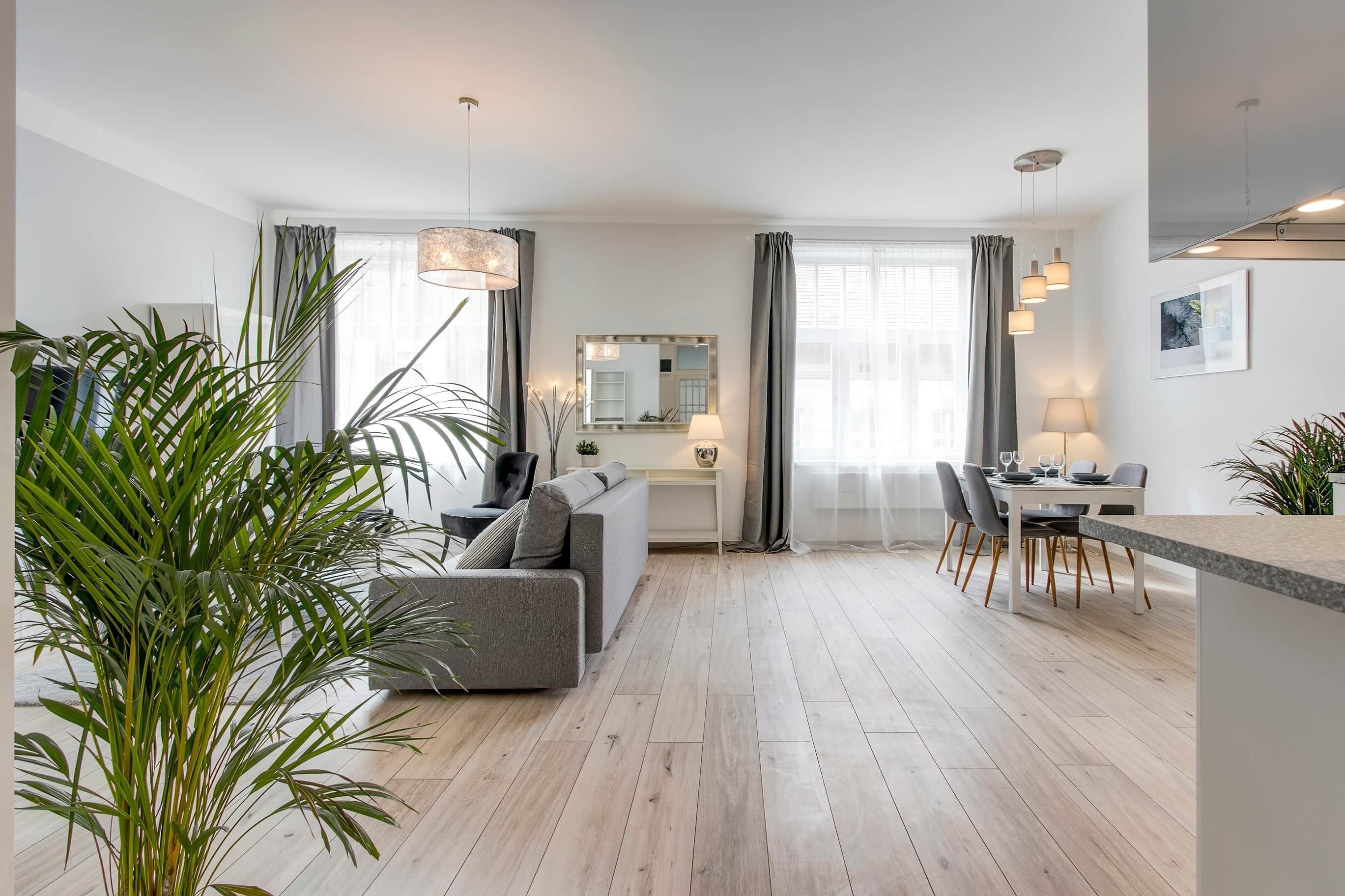 A modern living room with a grey sofa, a dining table with chairs, and a kitchen area with a hanging light fixture.