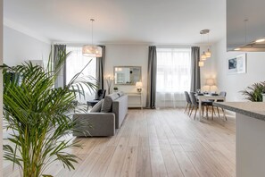 A modern living room with a grey sofa, a dining table with chairs, and a kitchen area with a hanging light fixture. - MidTerm - Rózsa Apartments - Daniel (Budapest)