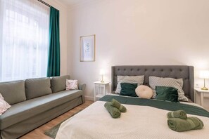 A hotel room with a bed, a sofa, a bedside table, and a window with curtains. - IMCT - Rose Apartment (Budapest)