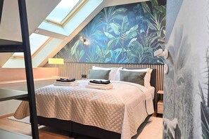 A bedroom with a sloped ceiling, a bed with a quilted bedspread, a nightstand with a lamp, and a wall with a tropical leaf pattern. - IMCT - Urban Jungle Apartments 04 (Budapest)