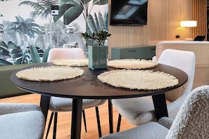 A round table with woven placemats, a potted plant, and grey upholstered chairs in a room with a tropical mural and a television mounted on the wall. - IMCT - Urban Jungle Apartments 04 (Budapest)