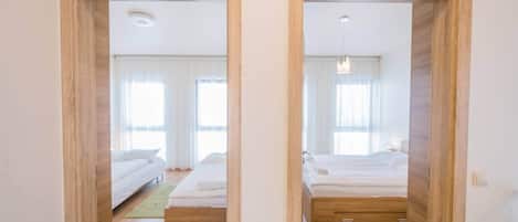 Una habitación de hotel con piso de madera, una cama con ropa blanca, una mesita de noche y una ventana con cortinas.