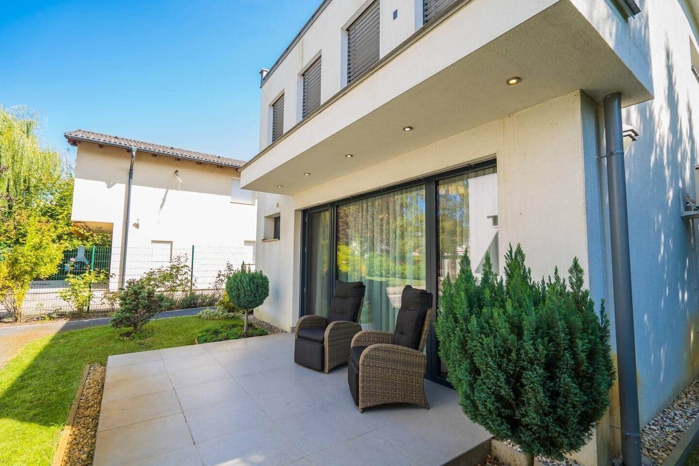 Una casa moderna con un patio, dos sillas de mimbre y un árbol pequeño.