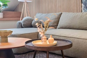 Ein modernes Wohnzimmer mit einem grauen Sofa, einem runden Couchtisch mit einer Vase getrockneter Blumen und einer kleinen Pflanze auf einem Beistelltisch.
