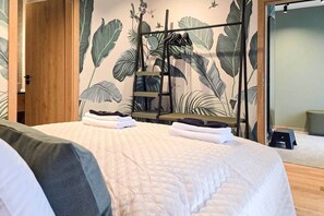 A neatly made bed with folded towels, a wooden door, and a wall with a large plant mural. - IMCT - Urban Jungle Apartments 03 (Budapest)