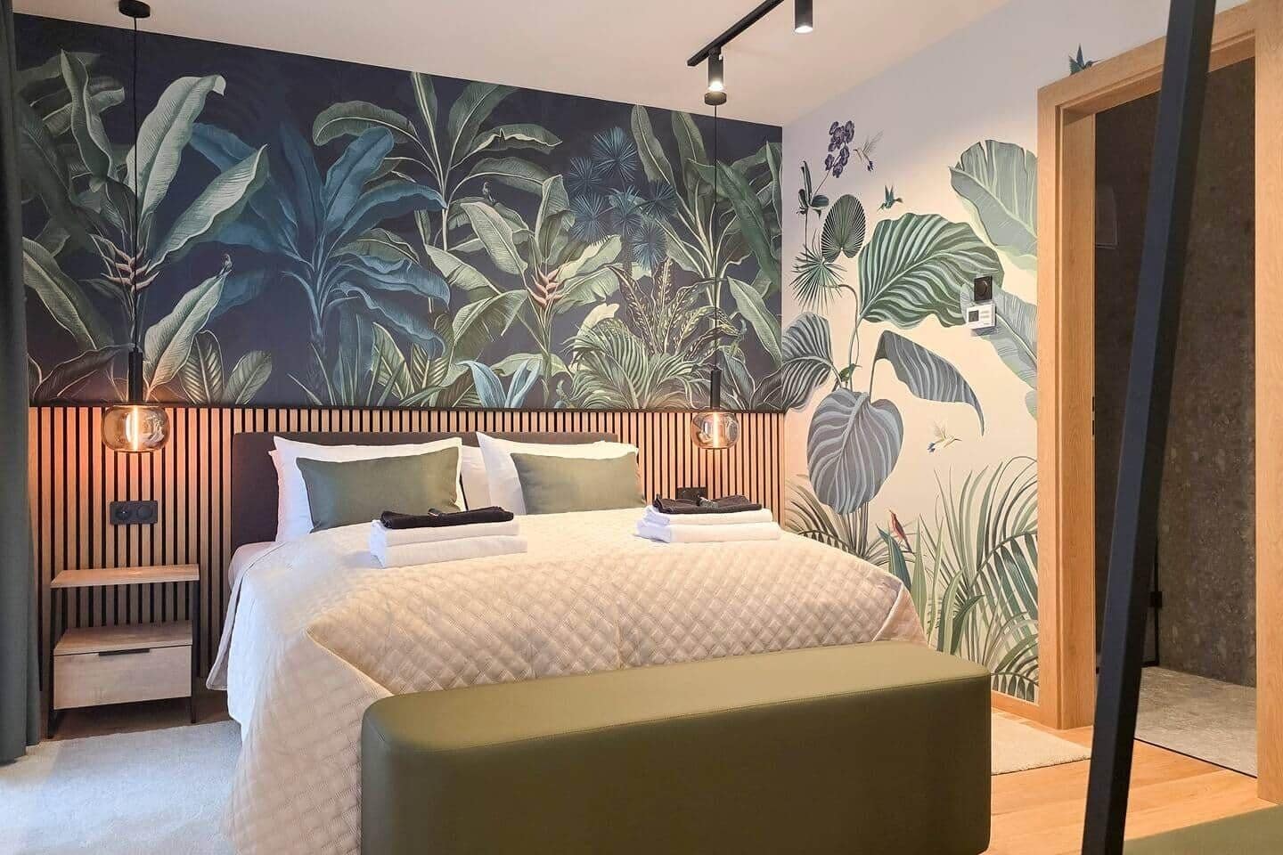 A bedroom with a large bed, a nightstand, a bedside table, and a wall mural of tropical foliage.