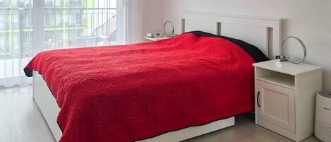 Una cama con una manta roja, una mesita de noche blanca con una lámpara y un armario blanco con un cubo de basura.
