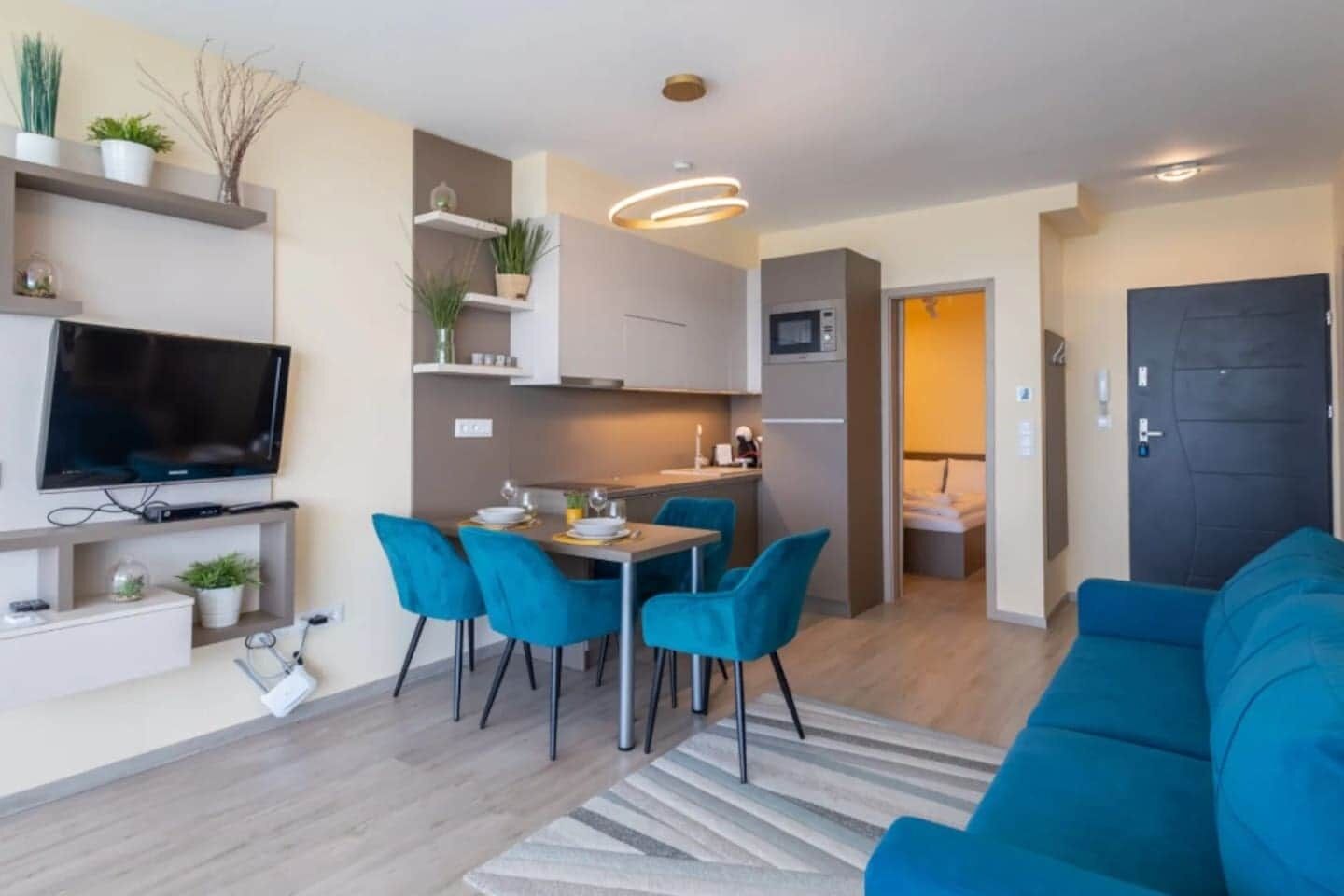 Ett modernt vardagsrum med en blå soffa, en matplats med blå stolar, en platt-TV och ett kök med mikrovågsugn.