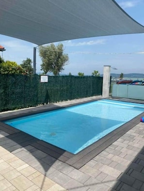 A rectangular swimming pool with a tiled deck, surrounded by a hedge and a covered area. - LB - LelleMarine Apartments A102 (Balatonlelle)