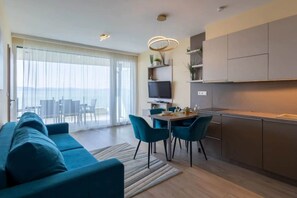 A modern living room with a sofa, dining table, and chairs, a kitchen area with cabinets, and a large window with a view. - LB - LelleMarine Apartments A102 (Balatonlelle)