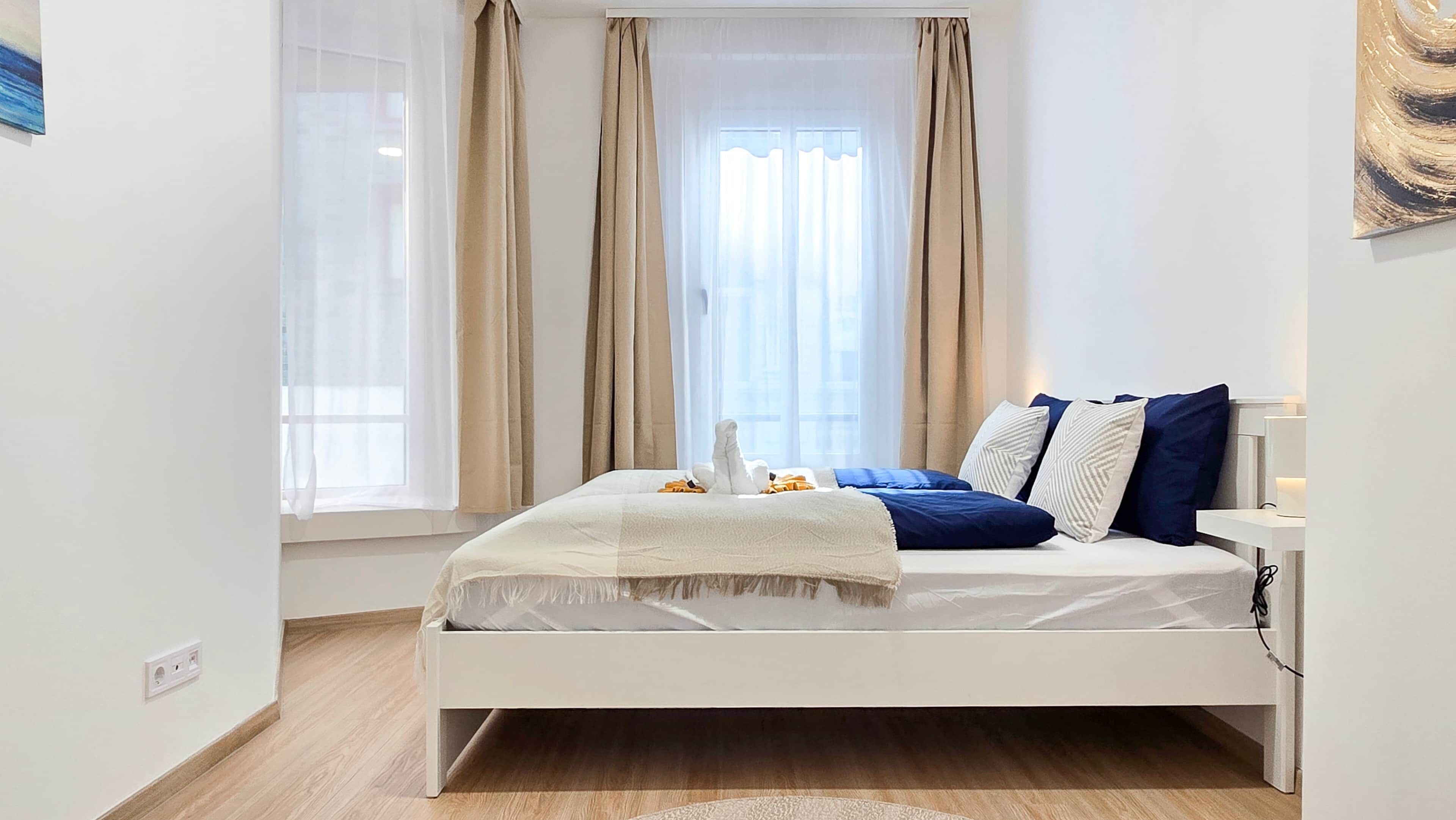 Une chambre moderne avec un lit blanc, des rideaux beige et une vue sur la ville.